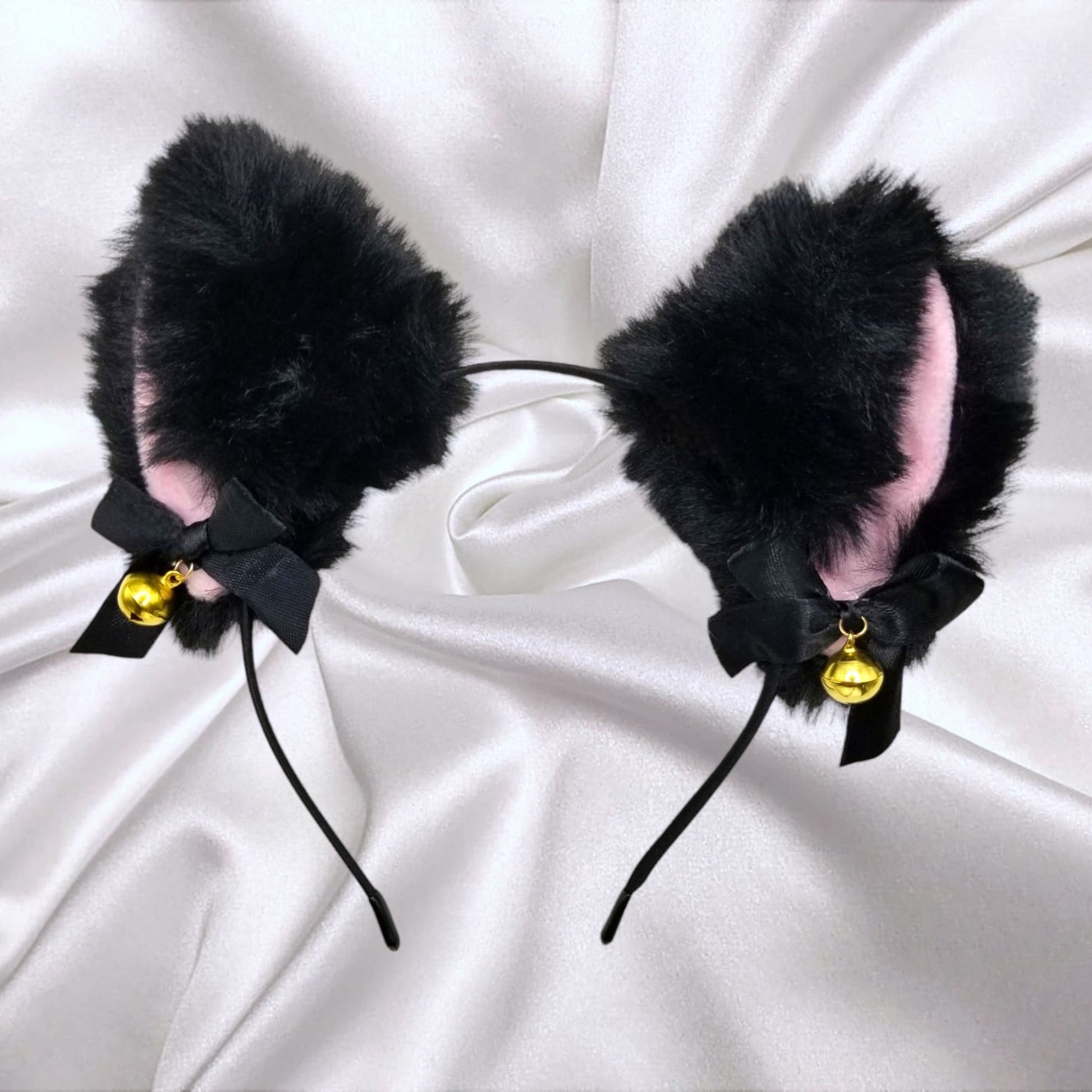 Black fluffy cat ears headband with pink inner lining, bow, and bell on satin background