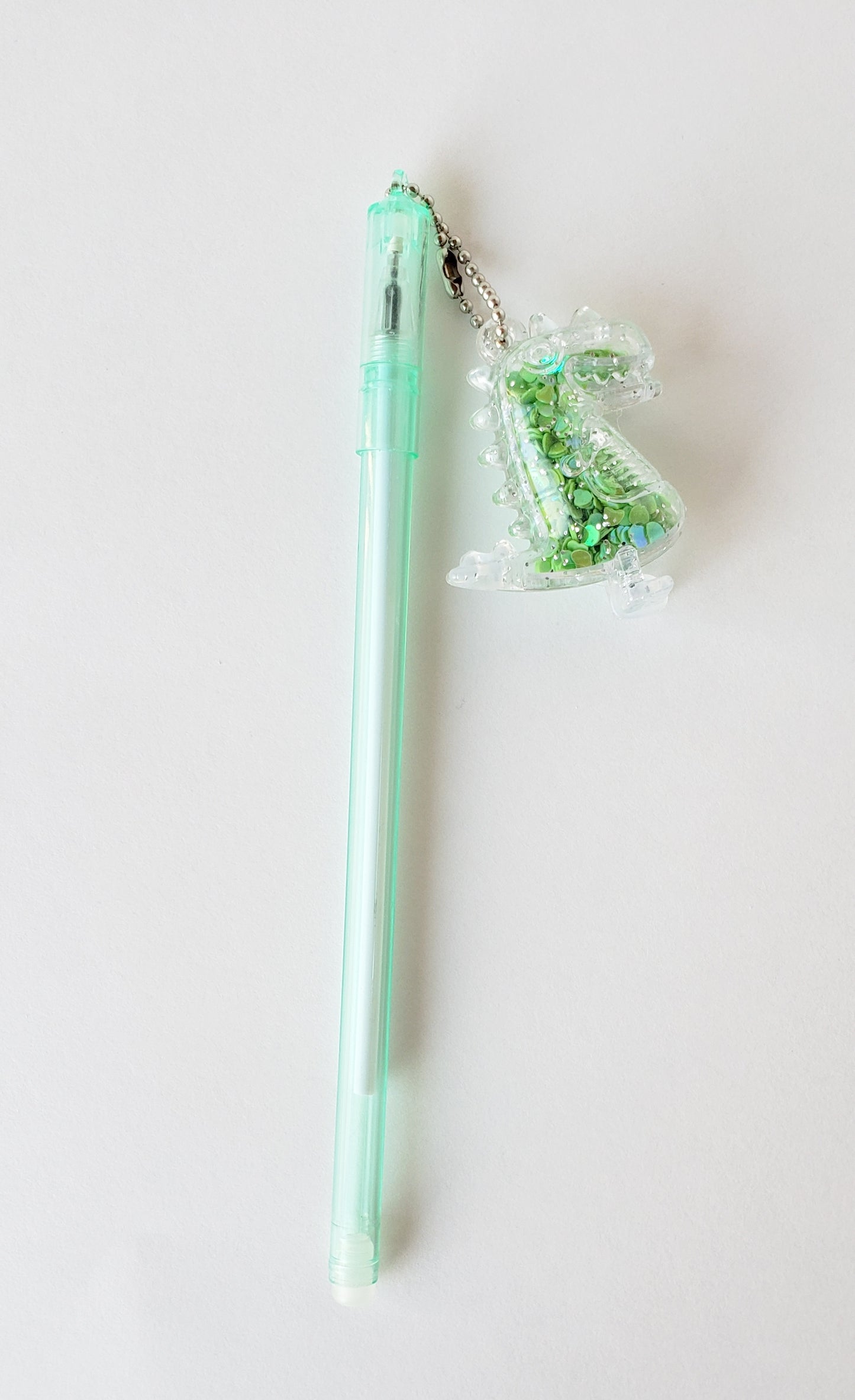 Flatlay of mint-green gel pen with attached silly dinosaur shaker charm on a white background