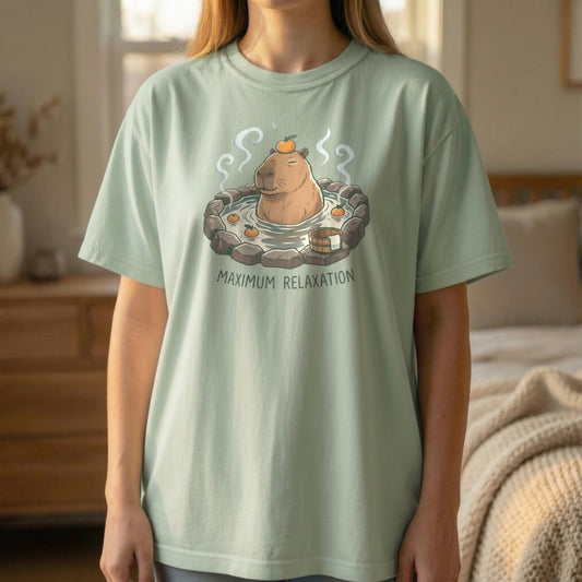 Model wearing a light green cottagecore t-shirt featuring a relaxed capybara hot spring illustration in a cozy indoor setting.