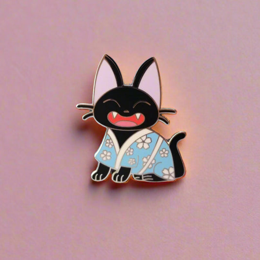 Flatlay of enamel pin featuring a smiling black cat wearing a light blue floral kimono on a pink background