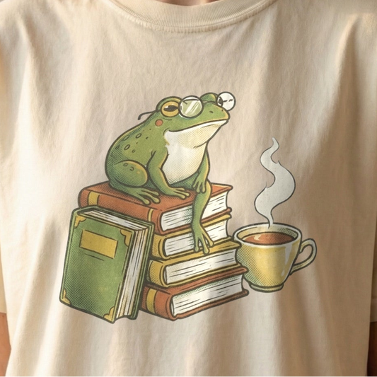 Front view of the Book Club Frog Tee in Ivory by Confetti Kitty. A vintage-style graphic of a green frog wearing round glasses perched on a stack of books next to a steaming cup of coffee, centered on the chest of a soft ivory t-shirt with the neckline visible.