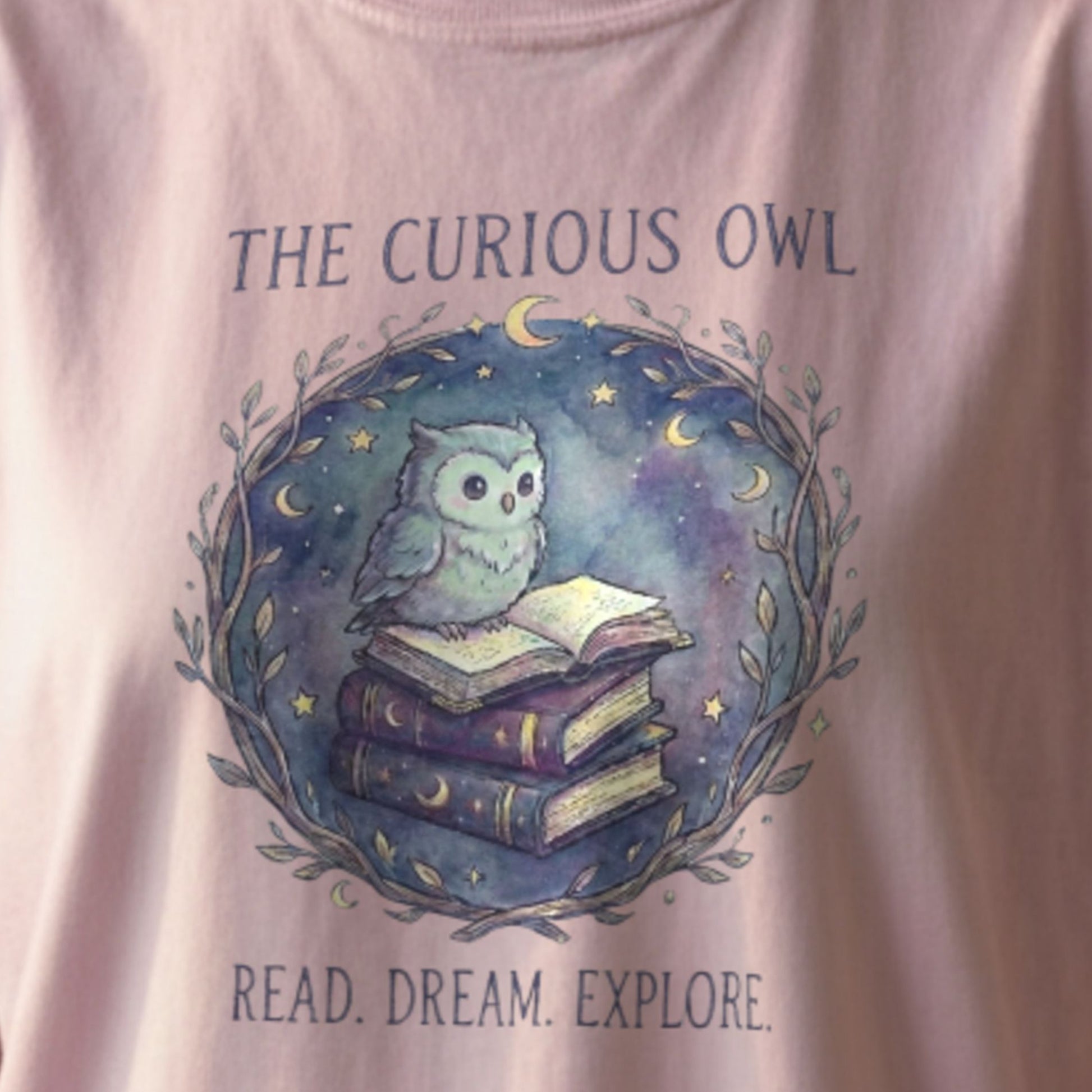 Close-up of the Curious Owl tee design showing a watercolor owl reading books surrounded by stars and moons on an orchid Comfort Colors shirt