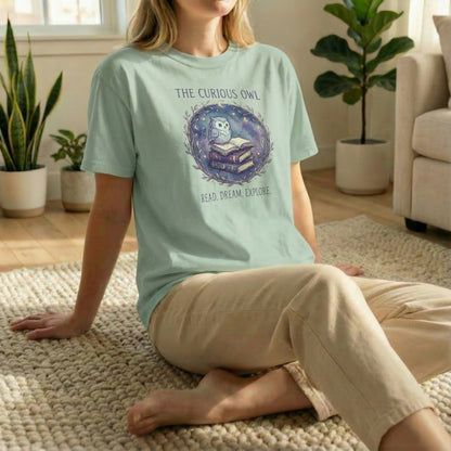Relaxed floor-seated lifestyle photo of the Curious Owl tee in sage with the owl and books illustration visible