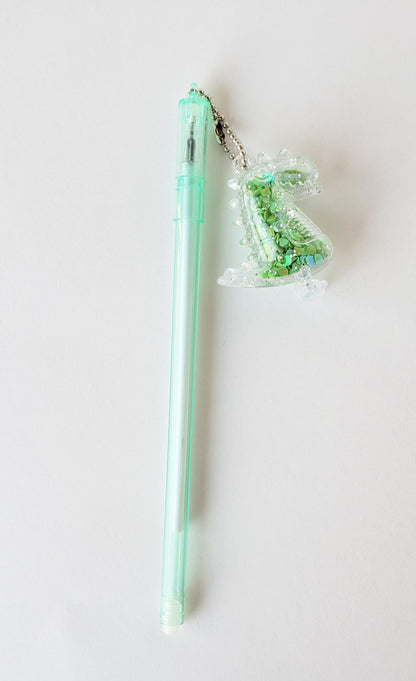 Flatlay of mint-green gel pen with attached silly dinosaur shaker charm on a white background