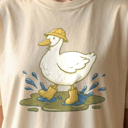 Duck in Rain Boots Tee on ivory Comfort Colors shirt featuring white duck wearing yellow rain boots and matching hat splashing in puddles, vintage cottagecore design.