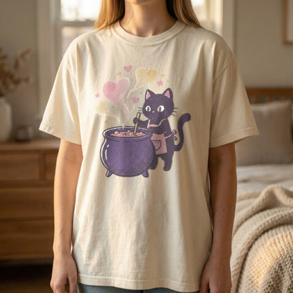 Mockup of a woman standing wearing an ivory t-shirt with a pixel art black cat in an apron stirring a purple cauldron.