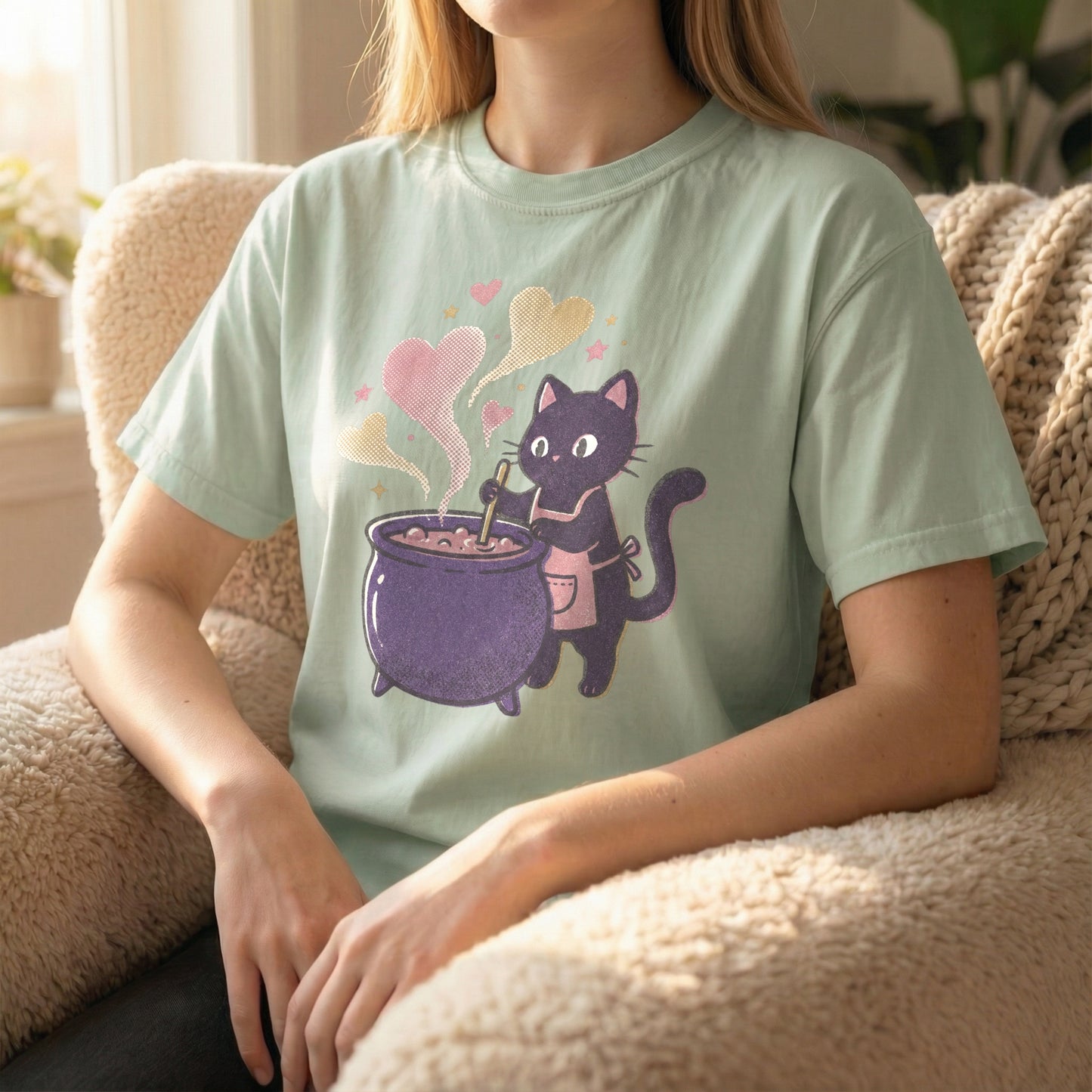 Woman sitting in a fuzzy chair wearing a light green t-shirt with a pixel art black cat and purple cauldron design.