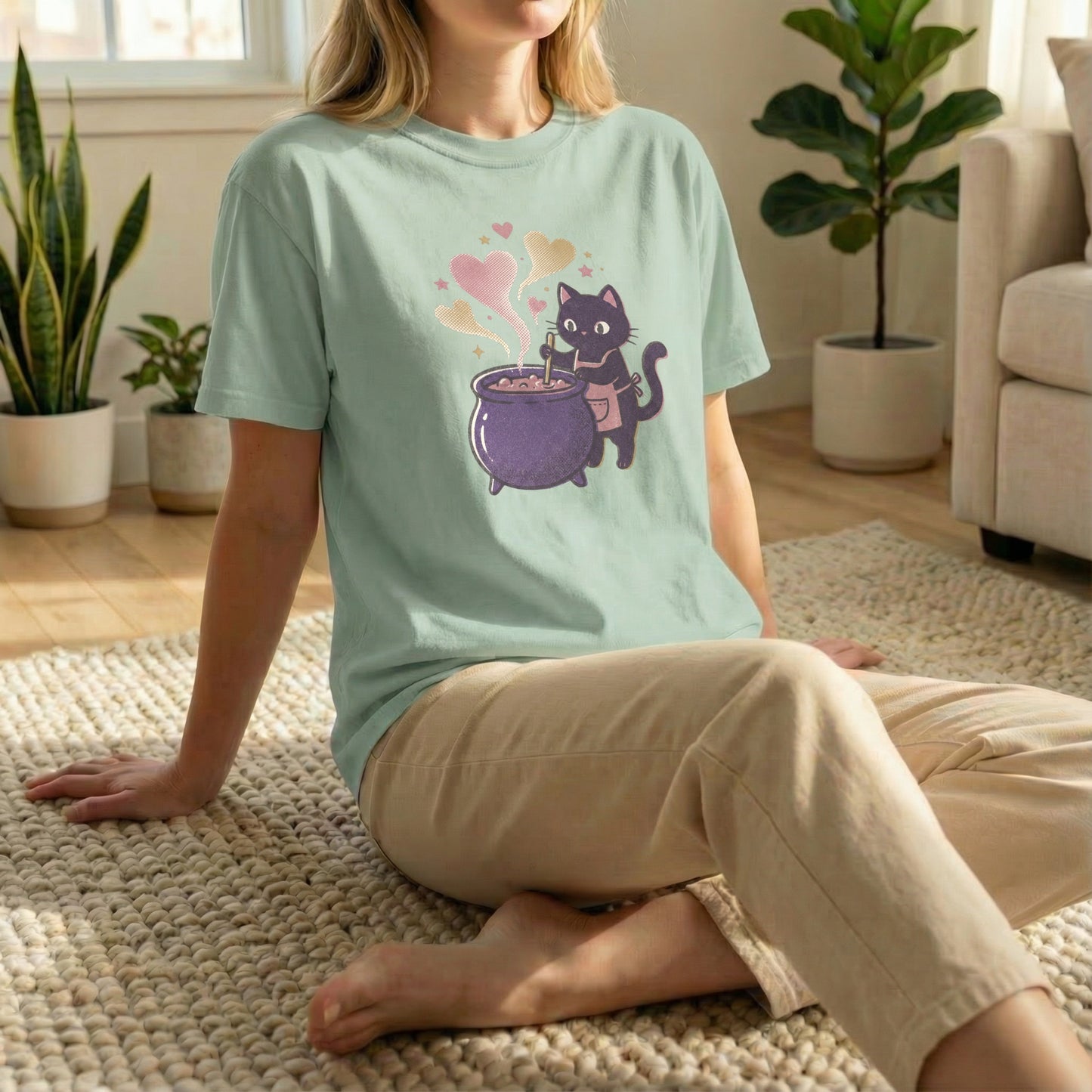 Woman sitting on a rug on the floor wearing a light green t-shirt with a pixel art black cat and purple cauldron design.