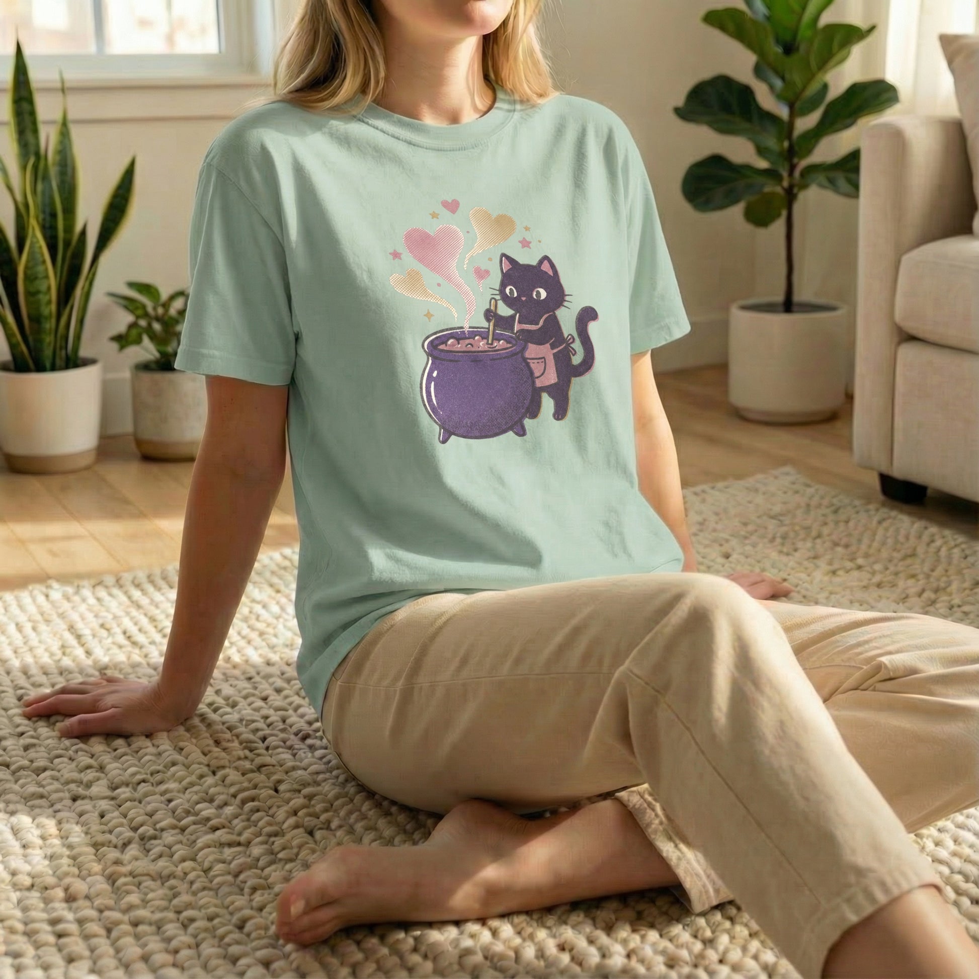 Woman sitting on a rug on the floor wearing a light green t-shirt with a pixel art black cat and purple cauldron design.