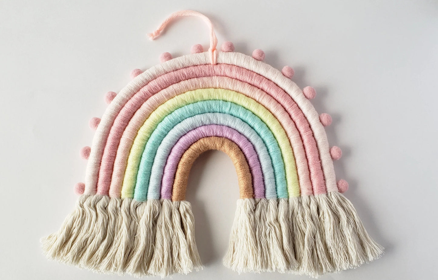 Flatlay of pastel rainbow wall decor showcasing hand-wrapped yarn arches in soft pink, yellow, mint, lavender, with gentle fringe tassels and a built-in hanging loop