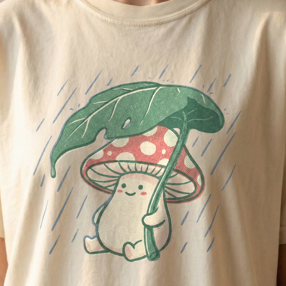 Rainy Day Mushroom Tee on ivory Comfort Colors shirt featuring smiling red and white toadstool holding green leaf umbrella in the rain, vintage cottagecore design.
