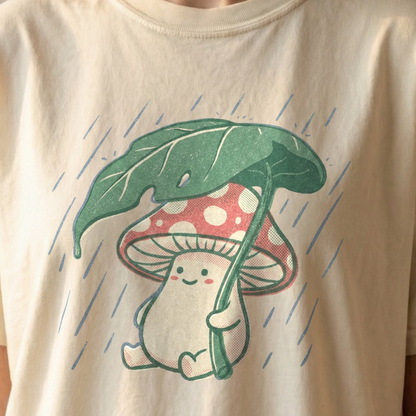 Rainy Day Mushroom Tee on ivory Comfort Colors shirt featuring smiling red and white toadstool holding green leaf umbrella in the rain, vintage cottagecore design.