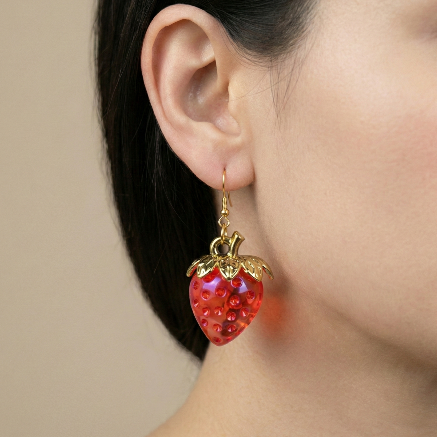Photorealistic close-up of a red, lightweight resin strawberry dangle earring with a gold-tone hook, worn by a model. The earring showcases the glossy finish and detailed fruit texture.