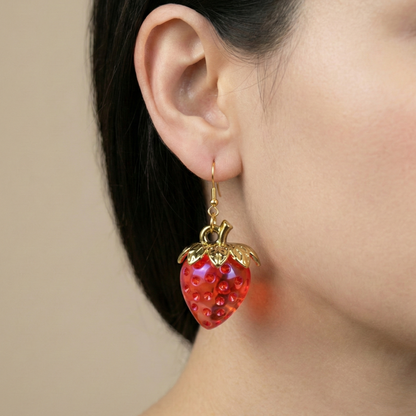 Photorealistic close-up of a red, lightweight resin strawberry dangle earring with a gold-tone hook, worn by a model. The earring showcases the glossy finish and detailed fruit texture.