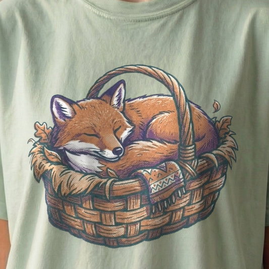 Sleeping Fox Tee on light green Comfort Colors shirt featuring vintage-style illustration of cozy fox curled up in woven basket, perfect for introverts and homebodies.
