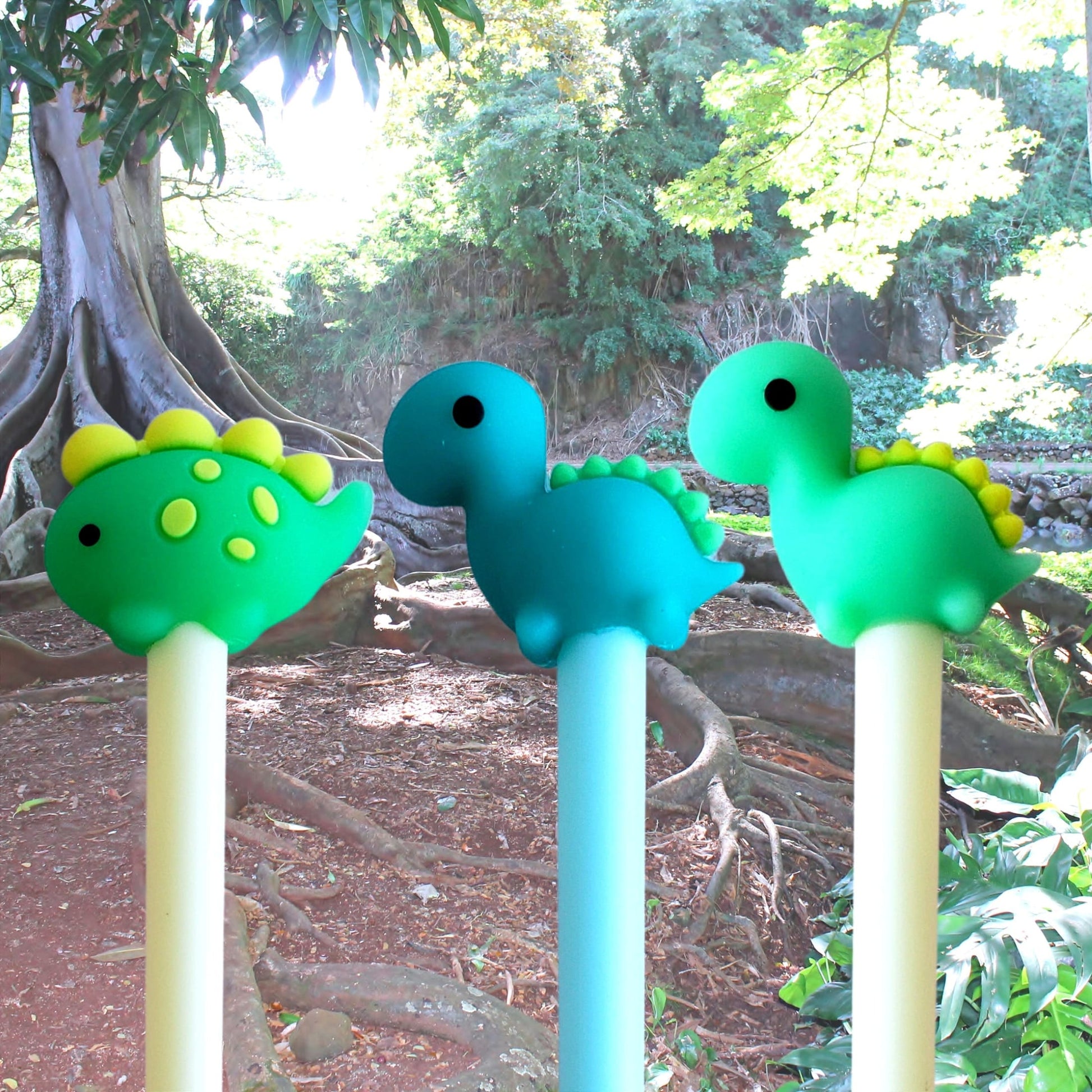 Three dinosaur pen toppers (green and teal) on pastel pens in an outdoor forest scene