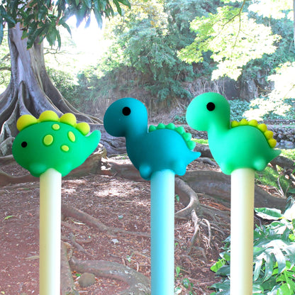 Three dinosaur pen toppers (green and teal) on pastel pens in an outdoor forest scene