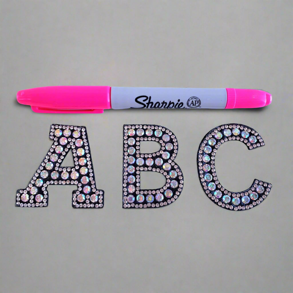 Three rhinestone letter patches spelling 'ABC' next to a pink Sharpie marker for size comparison.