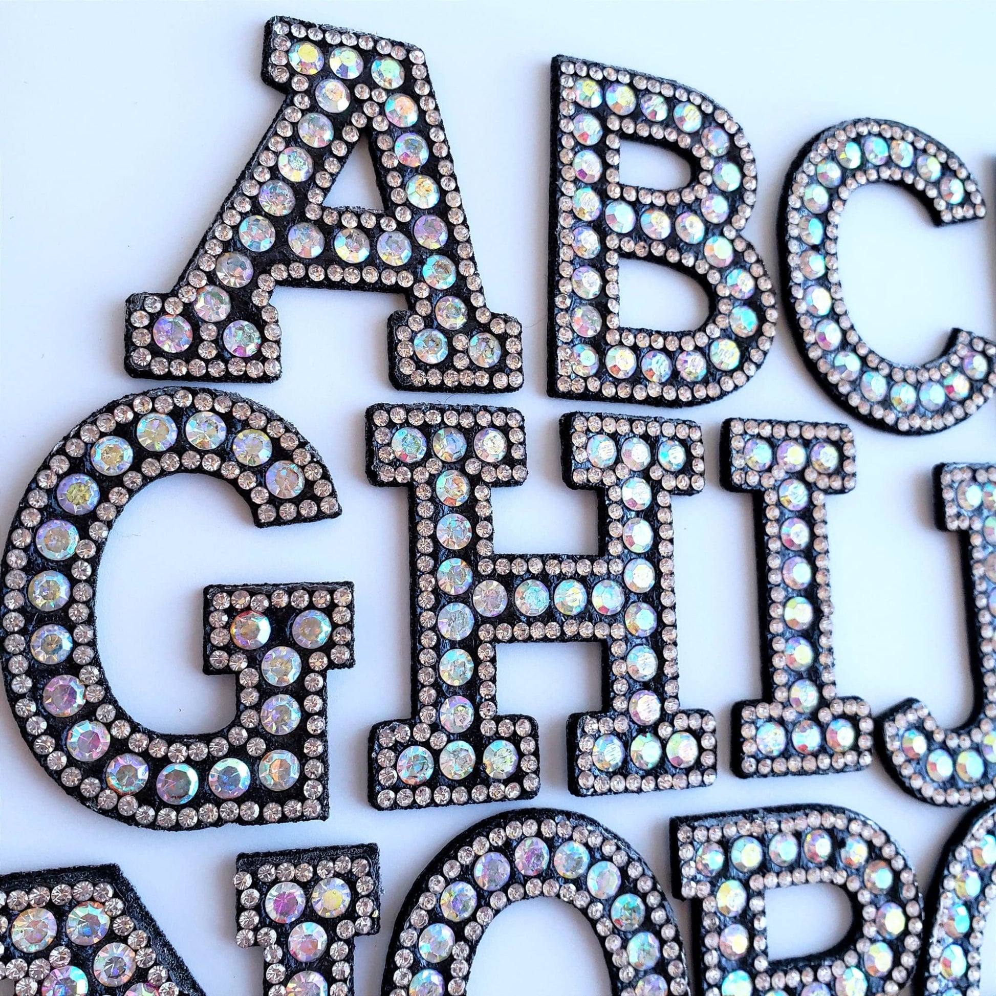 Close-up of sparkling rhinestone iron-on letter patches in the alphabet, featuring iridescent stones on a black base.